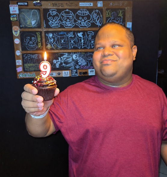 Diego Conceição sorri levemente enquanto segura um cupcake na mão direita. Sobre o bolo, há uma vela acesa no formato do número 9. 
