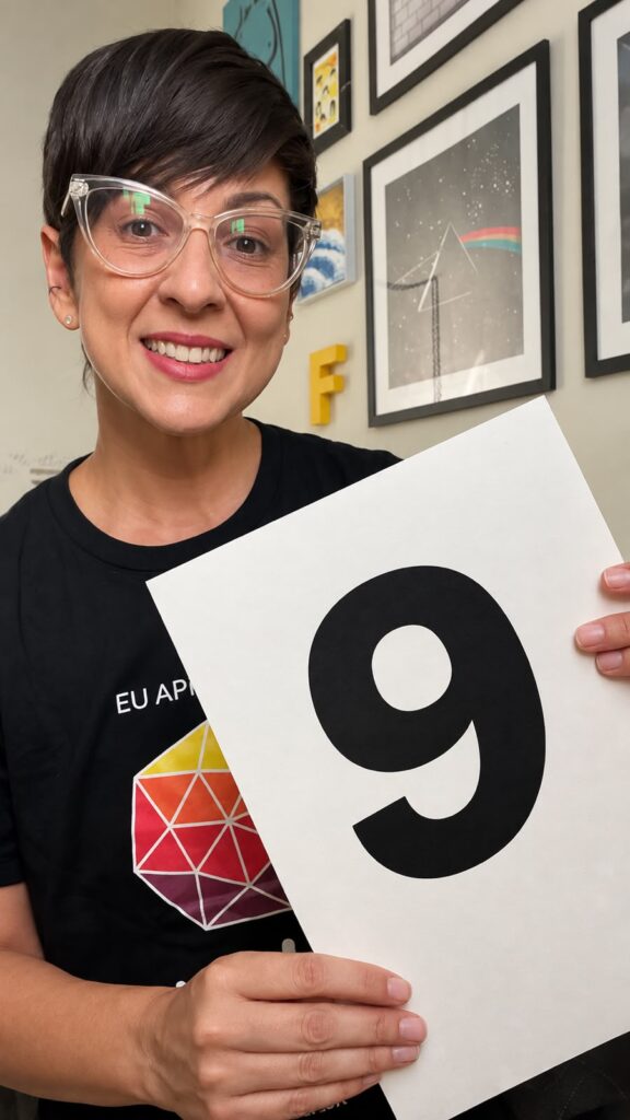 Fabiana Droppa sorri enquanto segura com as duas mãos uma folha de papel com o número 9 impresso. Ela veste uma camiseta preta com a marca do Web para Todos e a frase 'Eu apoio uma web mais inclusiva'. 
