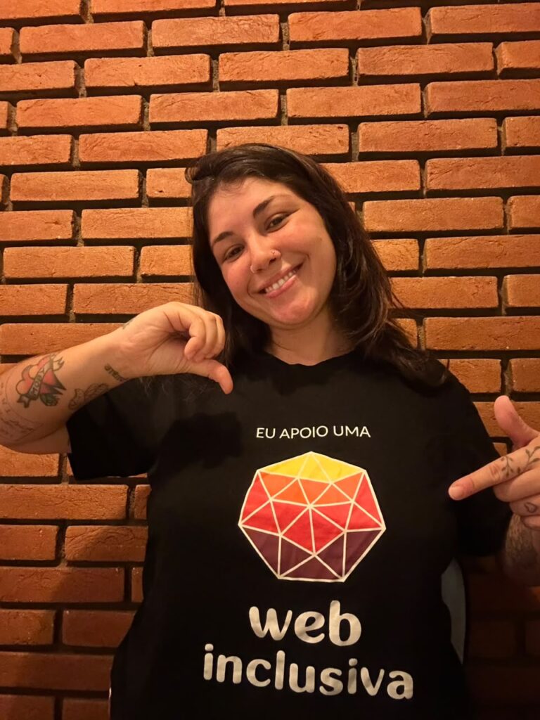 Gigi Villari sorri enquanto faz o sinal do número 9 em Libras com a mão direita e aponta, com o indicador esquerdo, para sua camiseta preta com a marca do Web para Todos e a frase 'Eu apoio uma web mais inclusiva'.
