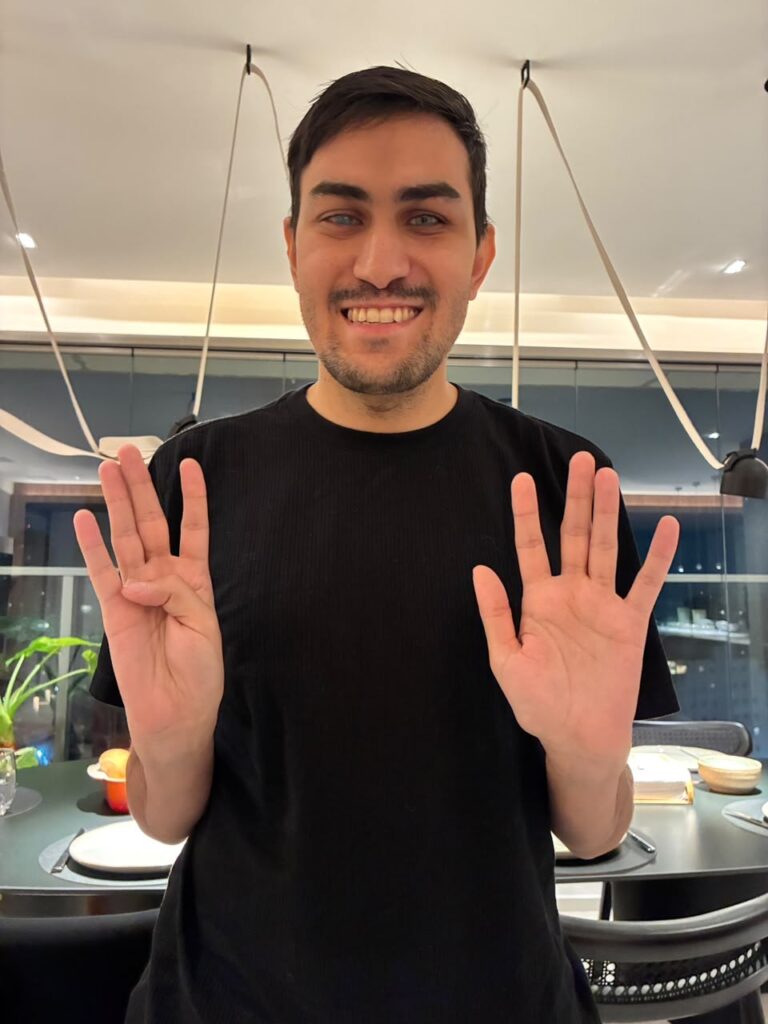 Gustavo Torniero sorri enquanto faz o número 4 com os dedos da mão direita e o número 5 com a mão esquerda, simbolizando o número 9.
