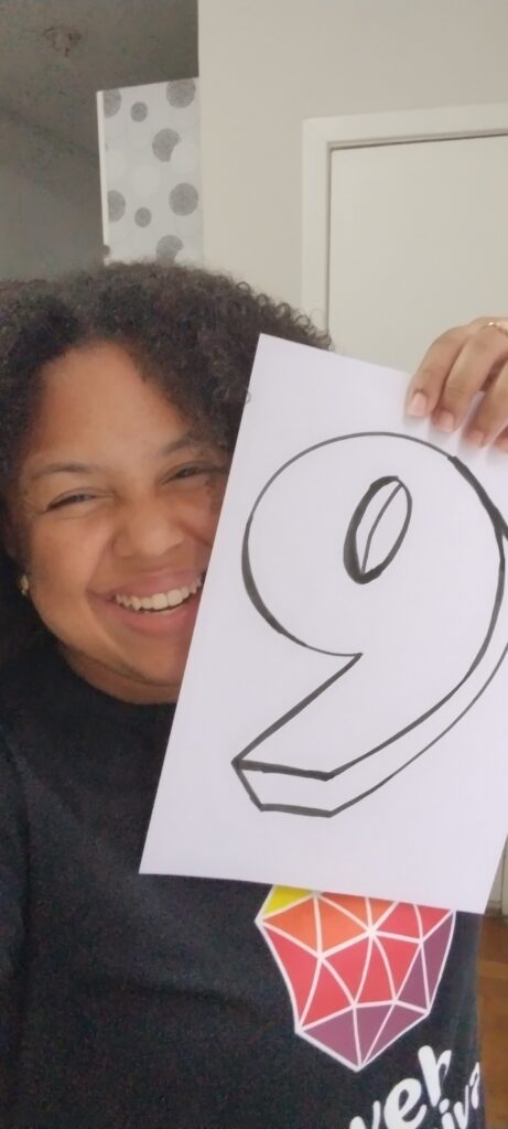 Íngrid Oliveira sorri enquanto segura, com a mão esquerda, uma folha de papel com o número 9 desenhado. Ela veste uma camiseta preta com a marca do Web para Todos e a frase 'Eu apoio uma web mais inclusiva'. 
