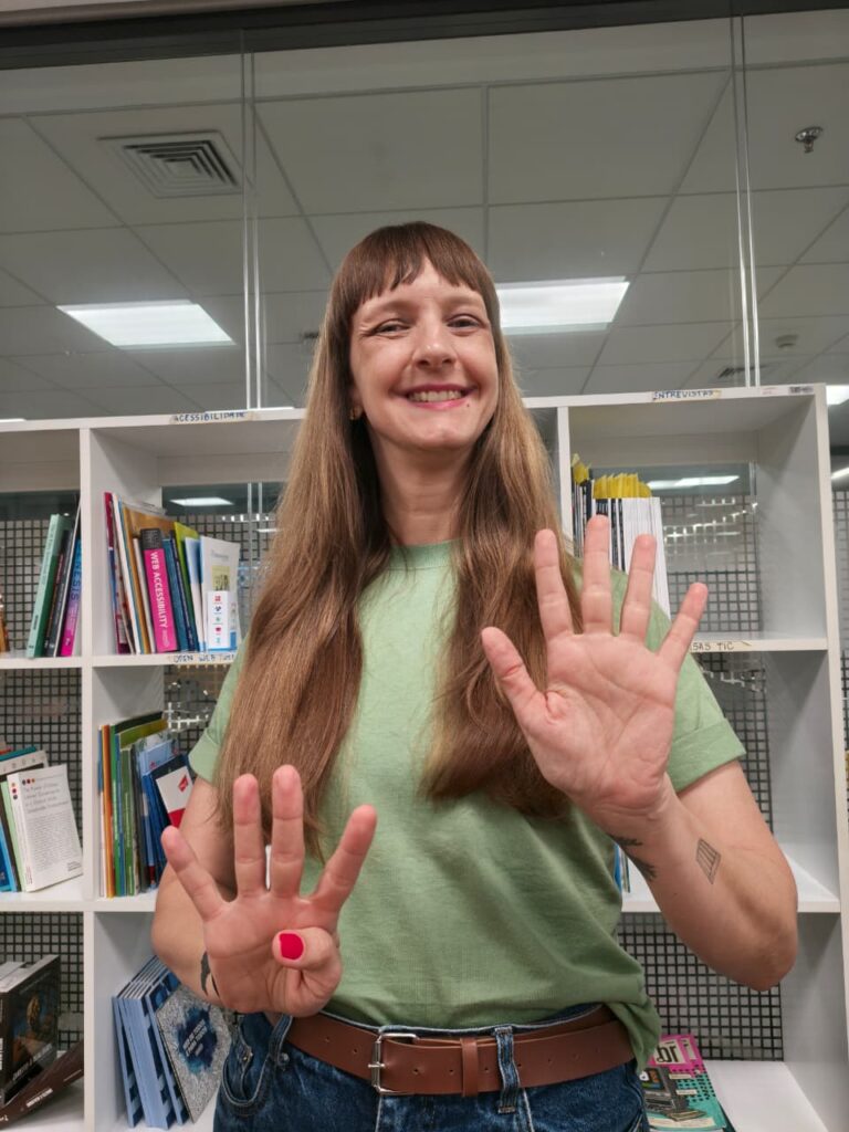 Jeniffer Deus sorri enquanto faz o número 4 com os dedos da mão direita e o número 5 com a mão esquerda, simbolizando o número 9.
