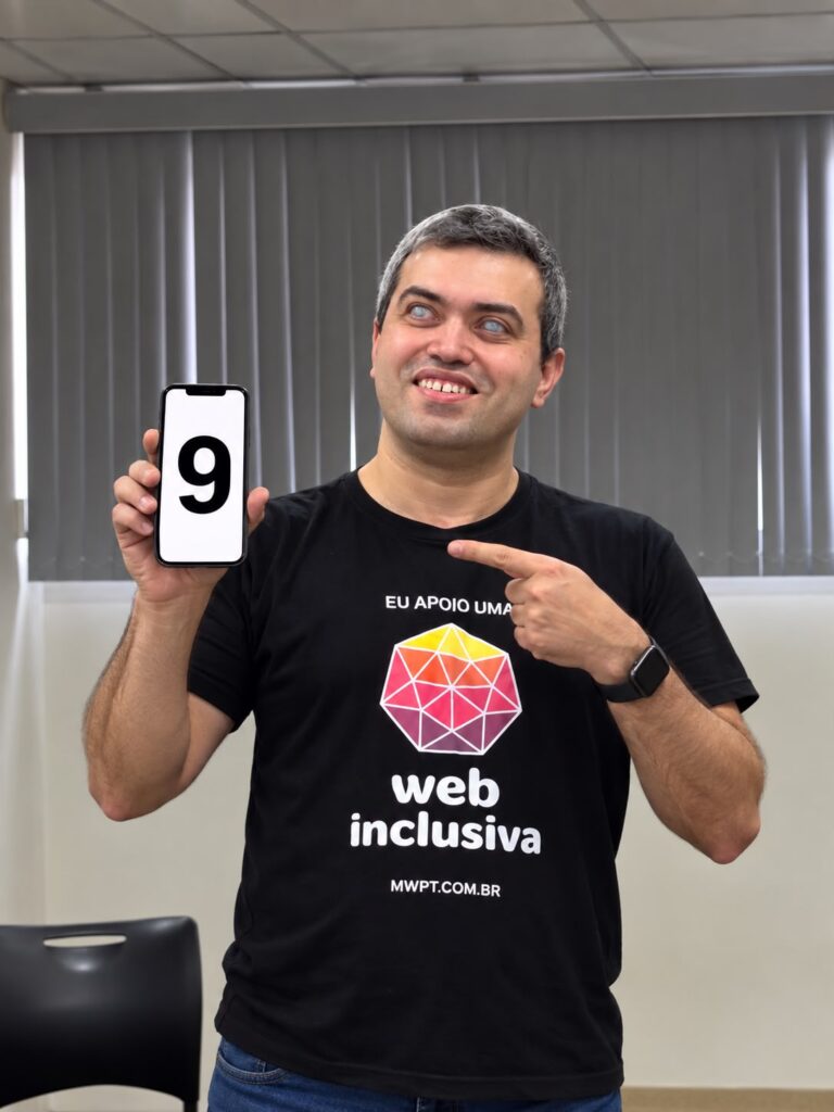 Leonardo Gleison sorri enquanto aponta para o celular na sua mão direita, que exibe o número 9 na tela. Ele veste uma camiseta preta com a marca do Web para Todos e a frase 'Eu apoio uma web mais inclusiva'. 
