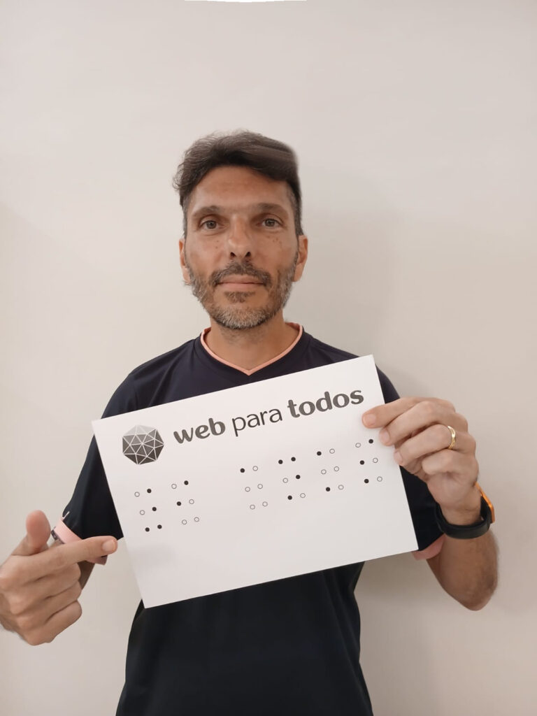 Marcelo Penha sorri levemente enquanto segura uma folha de papel com a mão esquerda e aponta com o indicador direito para ela. Na folha, há o logotipo do Web para Todos e o texto '9 anos' escrito em Braille logo abaixo. 
