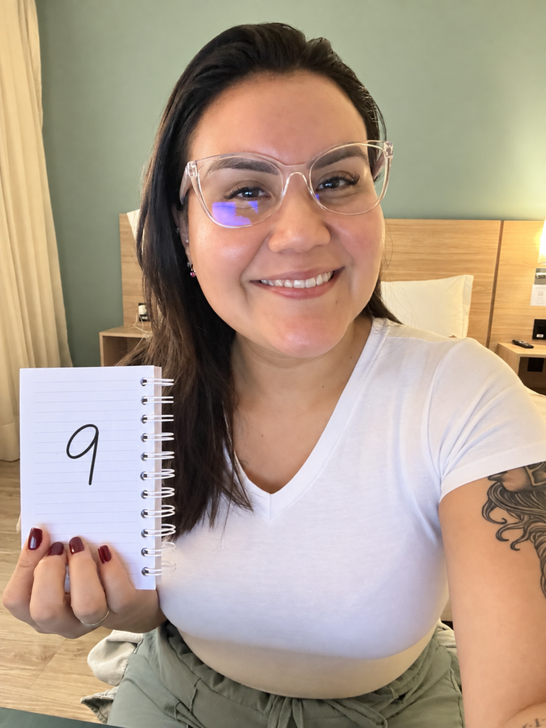 Natacsha Melo sorri enquanto segura com a mão direita um caderno aberto, que exibe o número 9 escrito em uma das páginas.