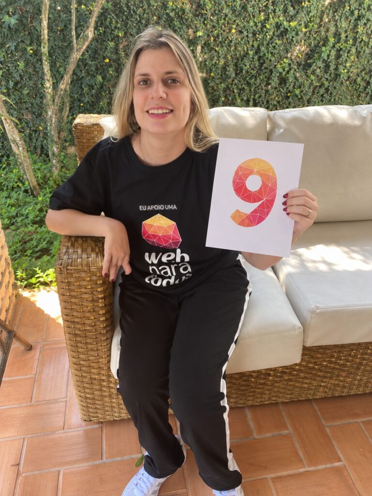 Nathalia Blagevitch sorri enquanto segura, com a mão esquerda, uma folha de papel com o número 9 desenhado. Ela está sentada em um sofá e veste uma camiseta preta com a marca do Web para Todos e a frase 'Eu apoio uma web mais inclusiva'.