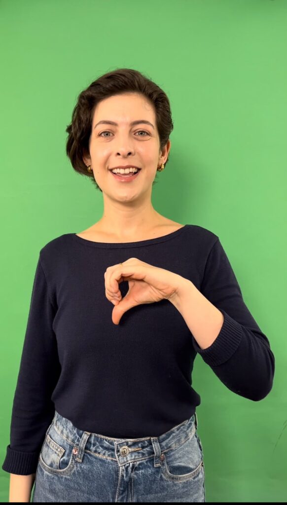 Paloma Bueno sorri enquanto faz o sinal do número 9 em Libras com a mão esquerda.