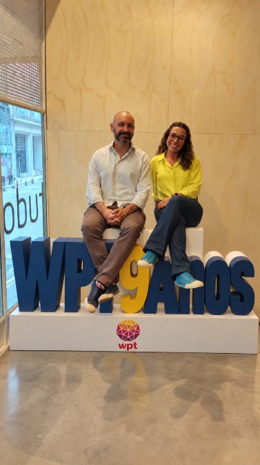 Rafael Públio e Aline Morais sorriem enquanto estão sentados em um banco. À frente deles, há um letreiro com a frase "WPT 9 anos" e no rodapé o logotipo do WPT. 
