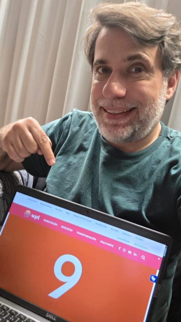 Reinaldo Ferraz sorri enquanto aponta, com o dedo indicador da mão direita, para a tela de um laptop. A tela exibe a página inicial do site do Web para Todos, com o número 9 destacado ao centro.
