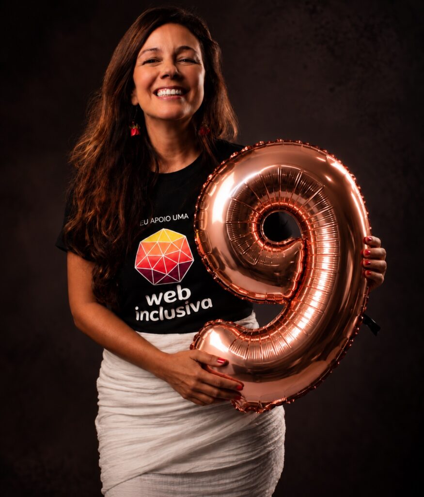 Foto de Simone Freire com um balão metálico no formato do numeral 9 nas mãos. Ela veste a camiseta com o logo do WPT com o texto "Eu apoio uma web mais inclusiva" e uma saia branca. Ela está maquiada e sorri.