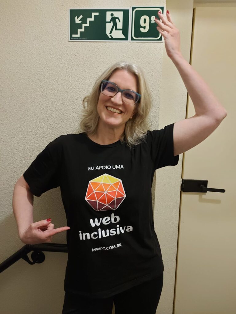 Suzeli Damaceno sorri em pé diante de uma parede, vestindo camiseta preta com a marca do Web para Todos e a frase 'Eu apoio uma web mais inclusiva'. Com a mão esquerda ela aponta para uma placa com o número 9 acima da sua cabeça, enquanto aponta para a camiseta com o indicador direito. 